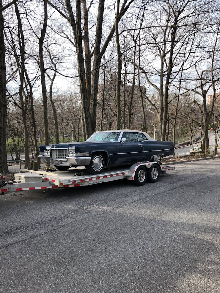 Classic blue car on a trailer amidst leafless trees on a street.